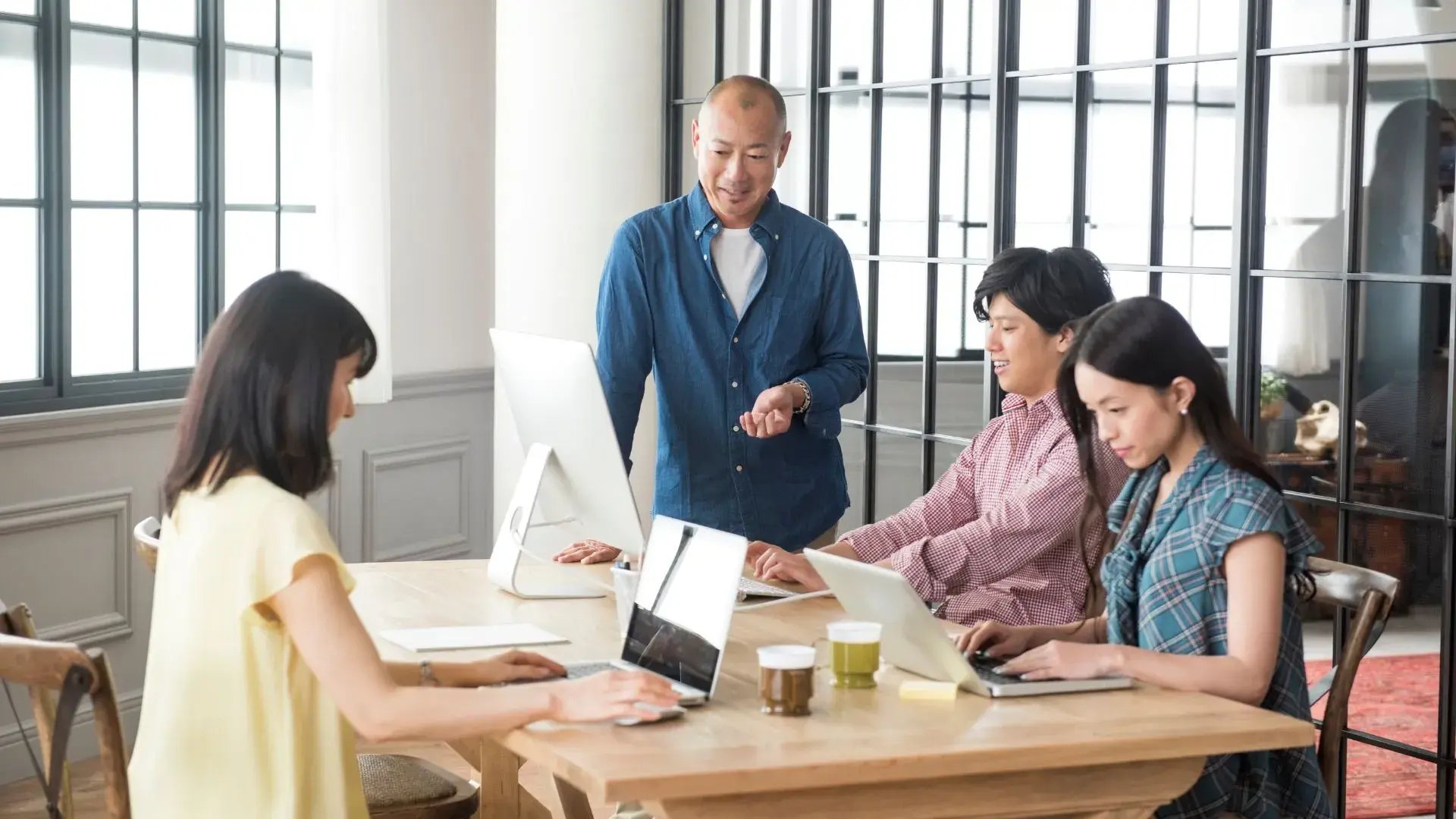 A group of four officemates are working together while facing their laptop screens. One of them seems to be explaining something while looking at one of the laptop screens.&nbsp;