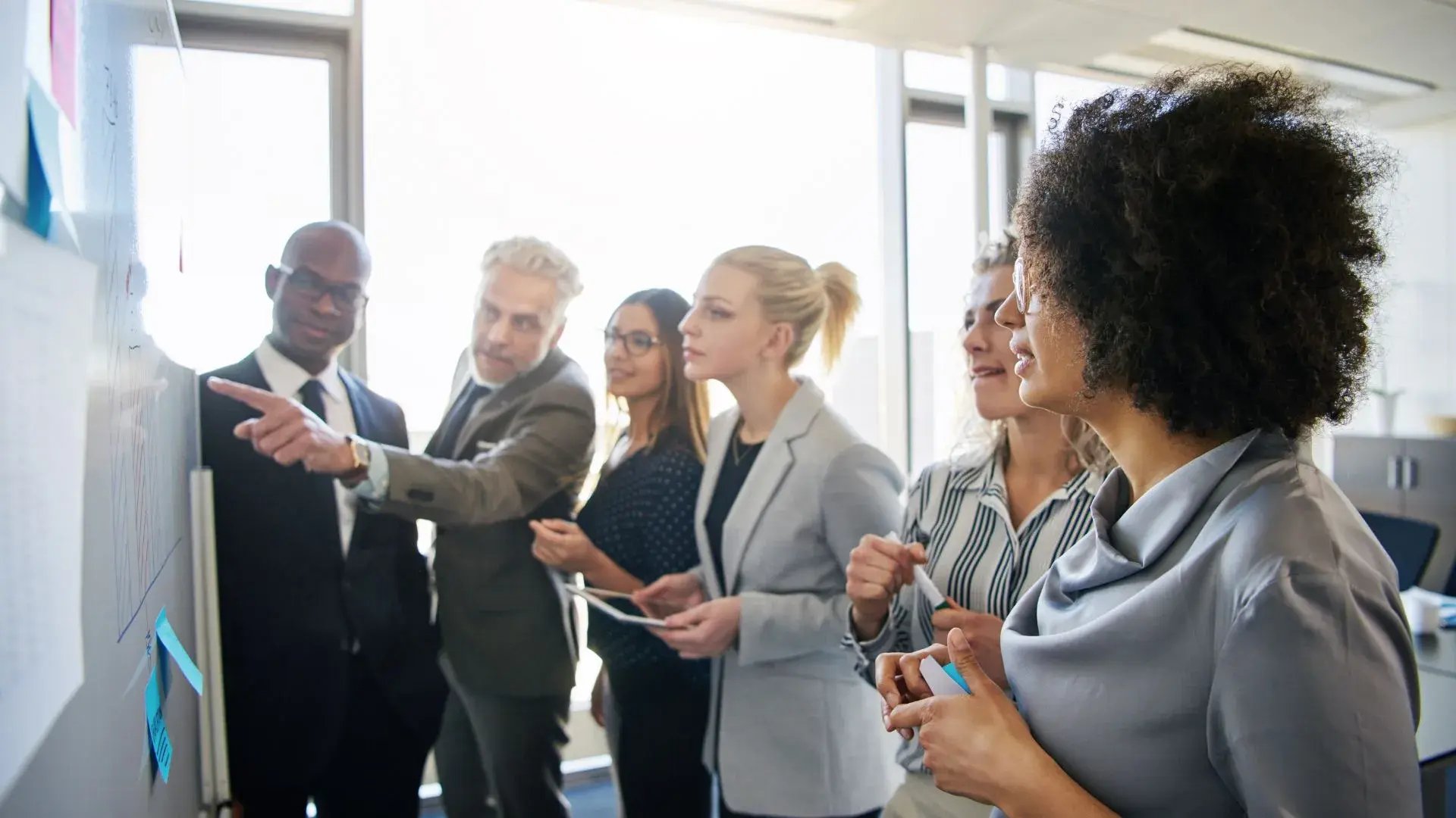 A team of corporate workers are gathered around a whiteboard filled with written notes. One of them is pointing to something on the board while the others are listening intently.&nbsp;