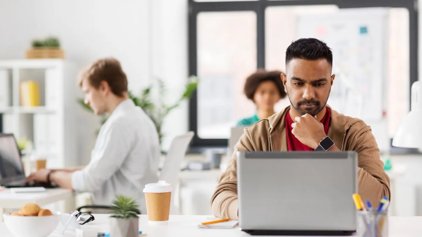 A male Indian office worker is in deep thought with his hand on his chin while staring at his laptop screen