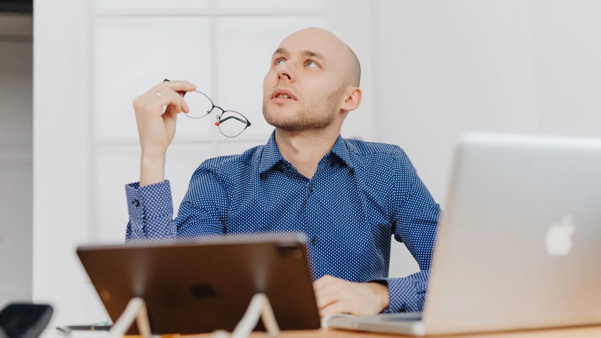 A bald man holding his eyeglasses while using his laptop