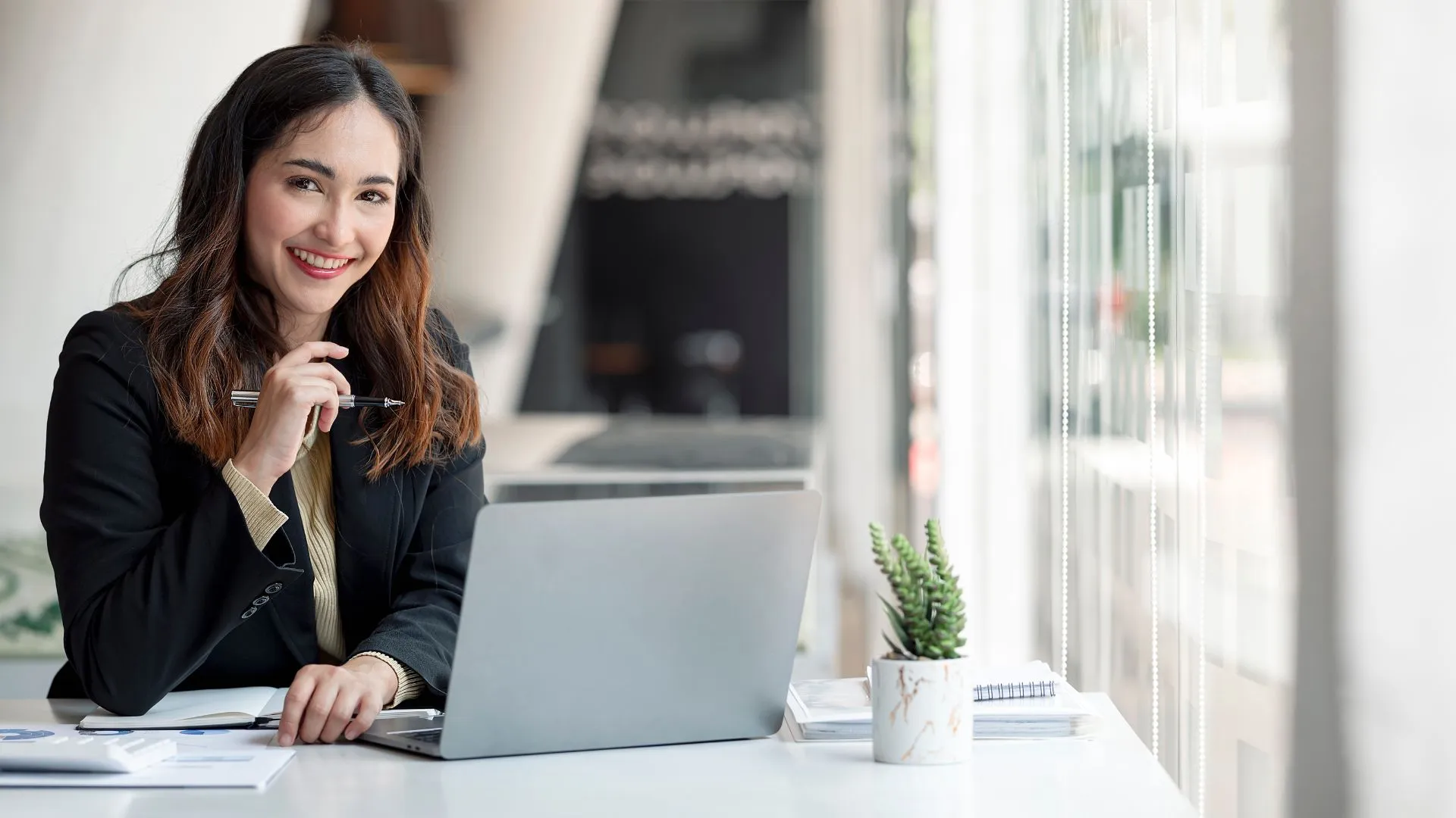 Alt image text:&nbsp; A woman in business attire sitting at a table, holding a pen with her laptop open and smiling at the camera