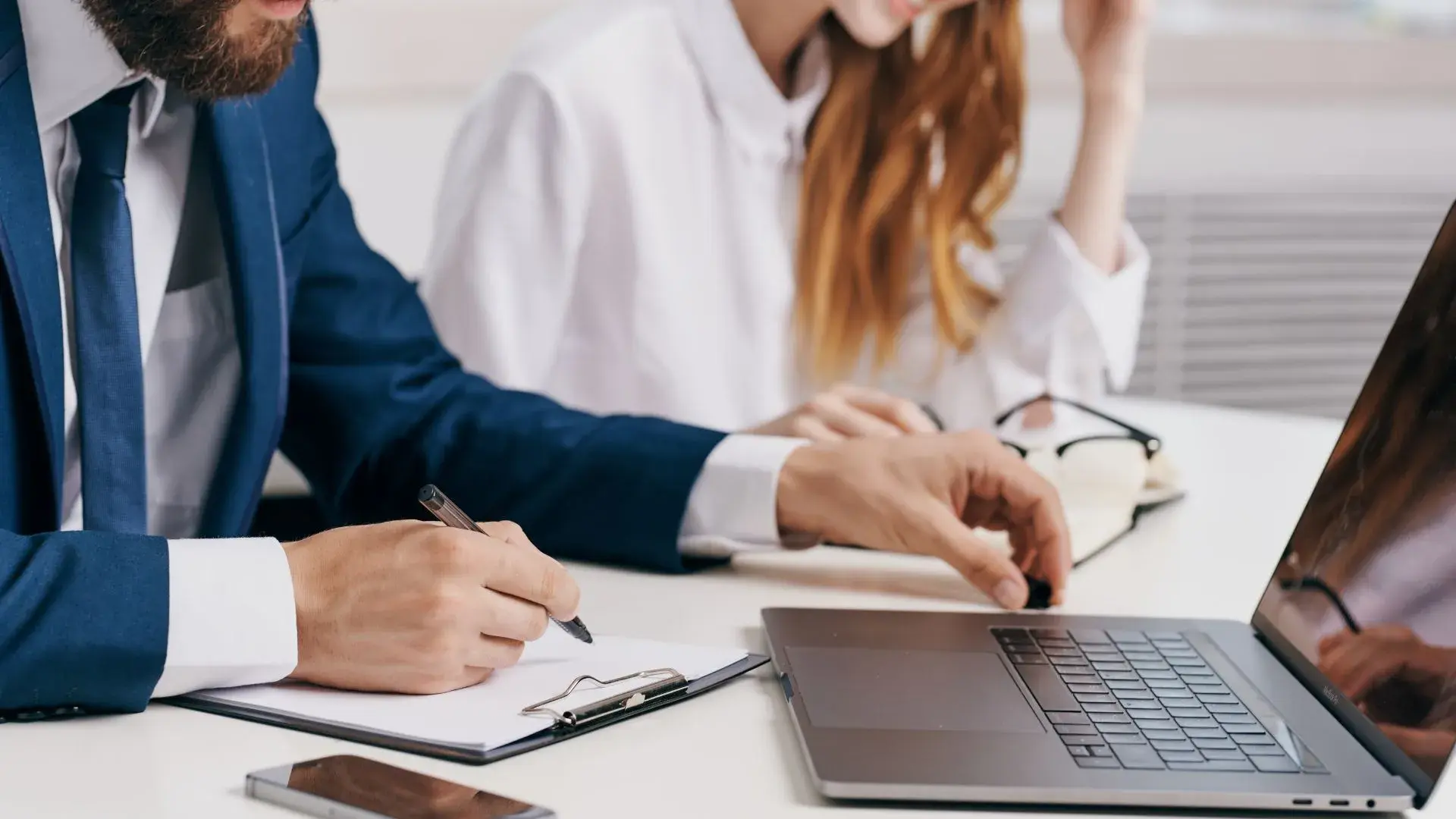 A male and a female office worker are working together. The male worker has his laptop open and is writing something on a clipboard. 